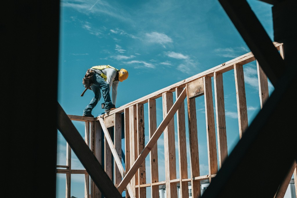 Construction worker in a hard hat and safety vest framing a house against a blue sky